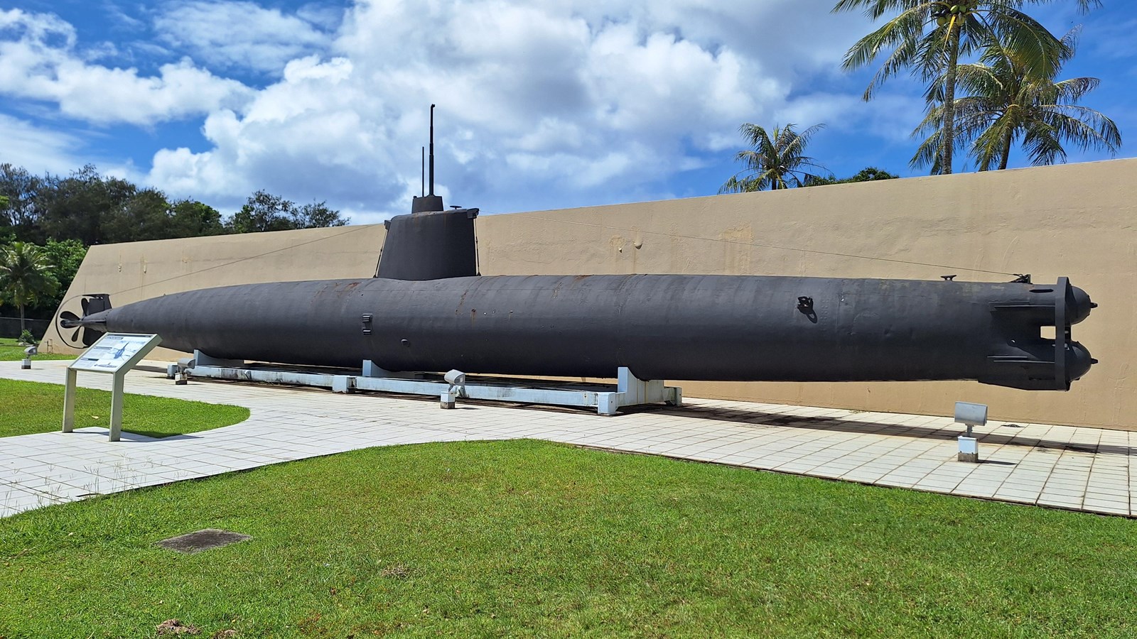 A large black submarine with a periscope resting on a concrete plinth in front of a concrete wall.
