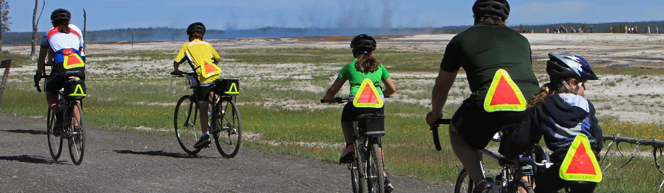 Five cyclists ride on a trail with steam rising from a geysers in the background.