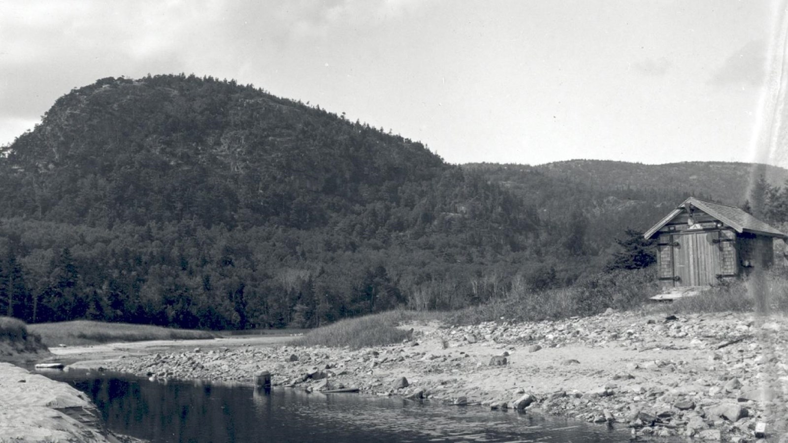 a small wooden building stands next to a sandy estuary in front of a mountain.