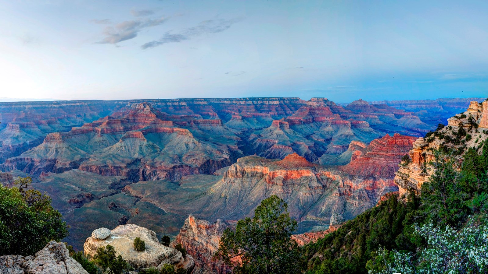 Under a pale blue sky, the landscape of Grand Canyon is illuminated by the fading light of sunset.