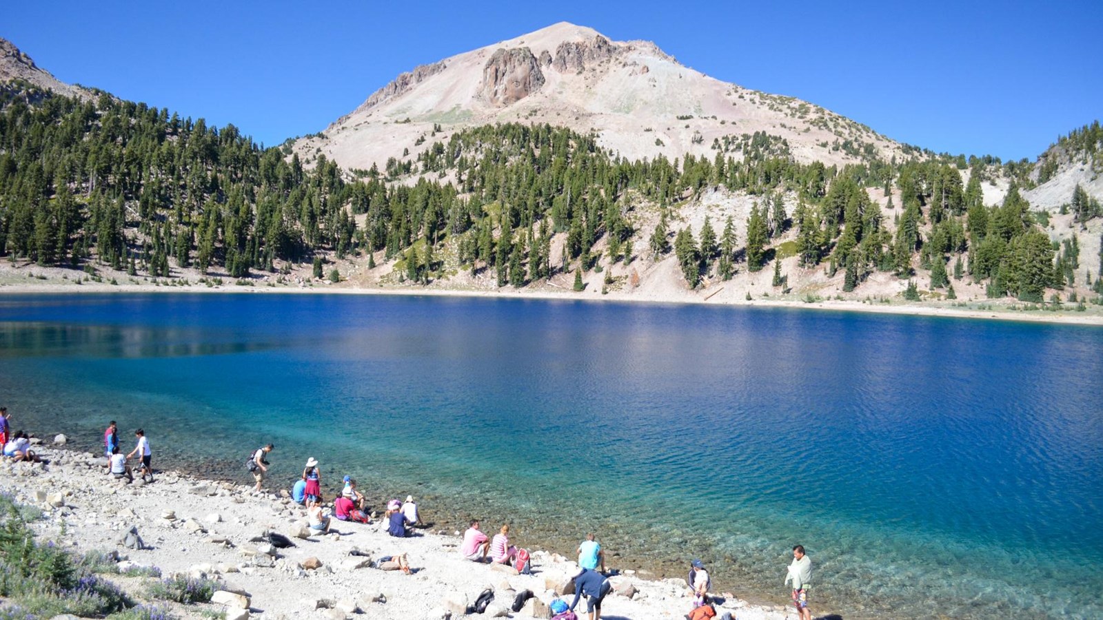 A dozen people spread out along the rocky shore of an alpine lake.