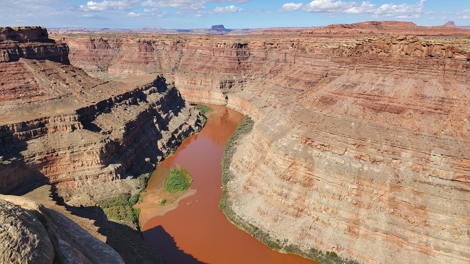 A chocolate-brown river cuts through a layered canyon on a bright, sunny day.