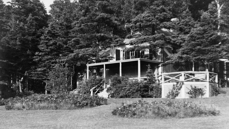 a large house with a covered porch and white trim sits in the shade of trees with a big lawn.