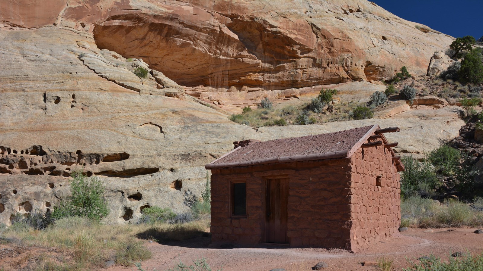 Small stone cabin with dirt roof, one door, one window, below cliffs.