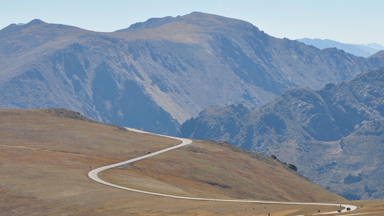 Vehicles are driving on Trail Ridge Road as the road snakes along tundra with mountains behind.