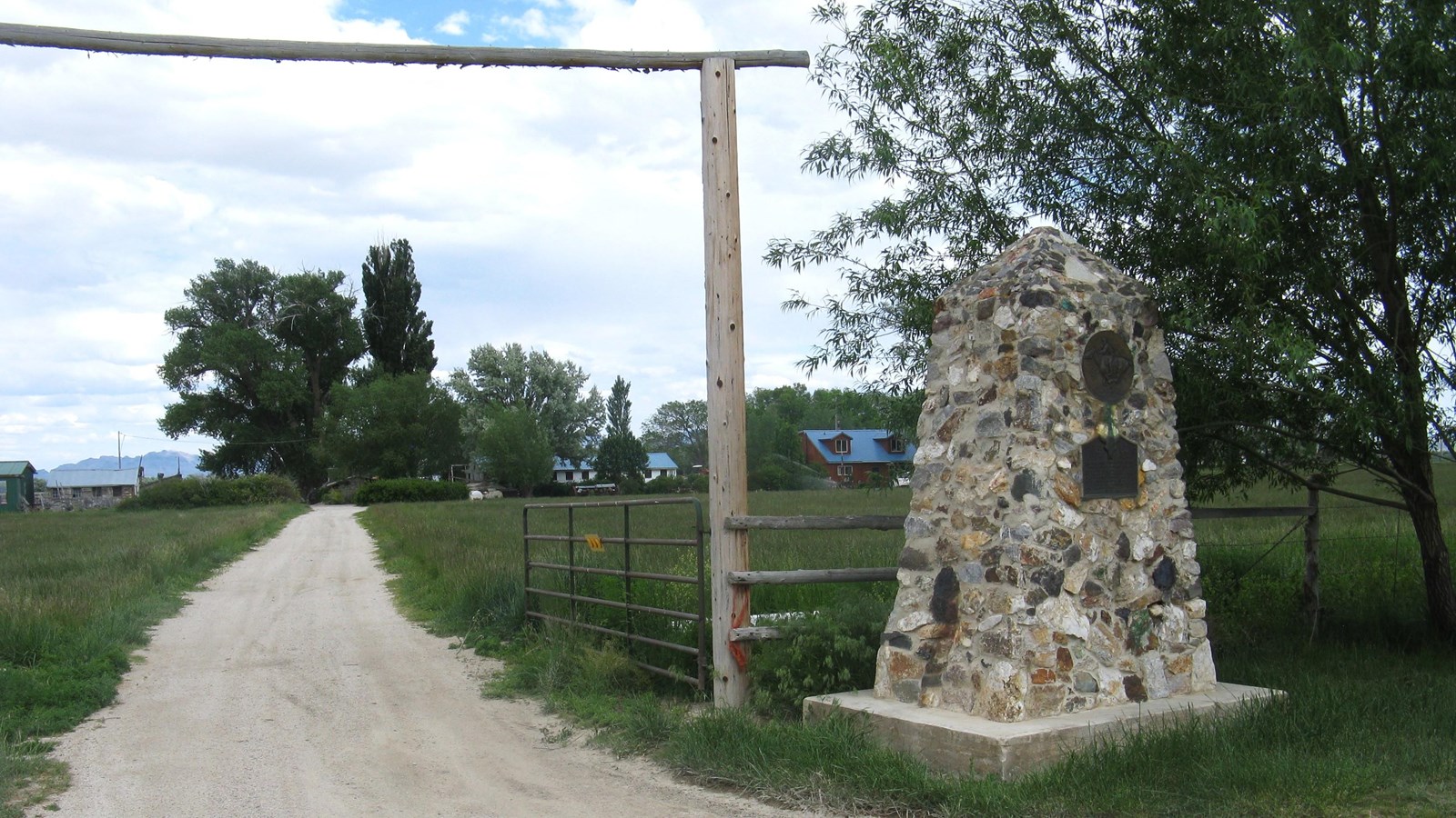 A stone obelisk sits to the right of a gravel driveway with grass and houses behind it.