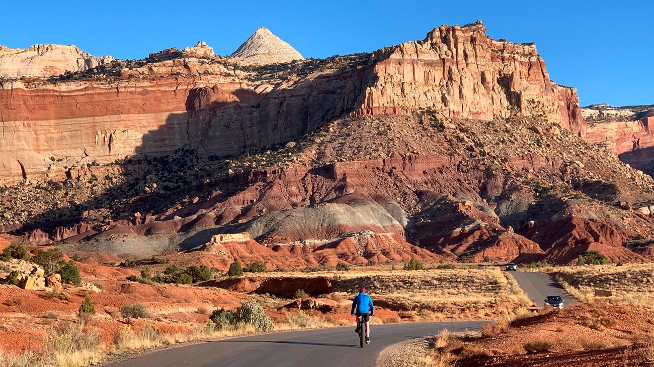 Person on bicycle on paved road with cars approaching and colorful cliffs above, and blue sky.