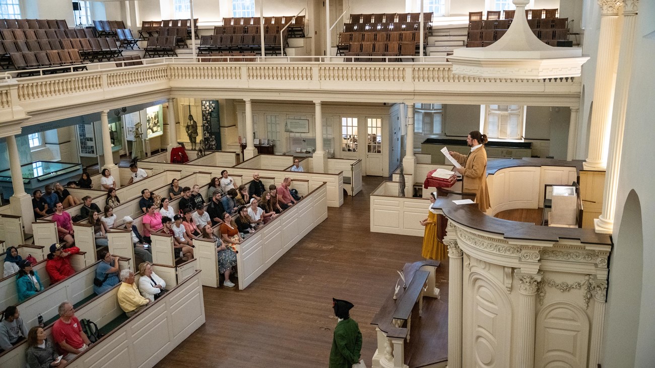 The two-story old south meeting house with people sitting in the rows of pews during a town meeting.