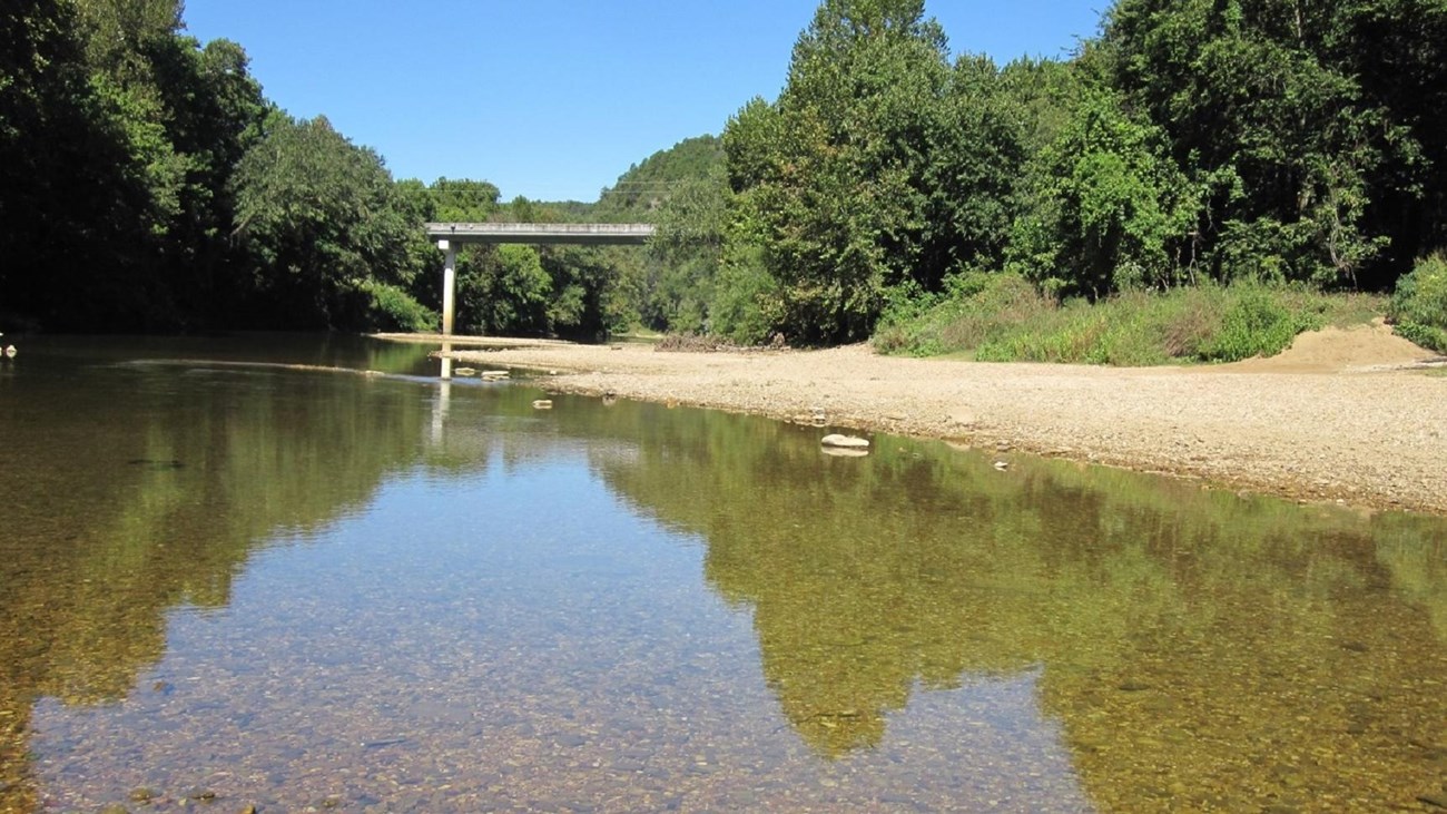 water in foreground with tan gravel at right and highway bridge in background