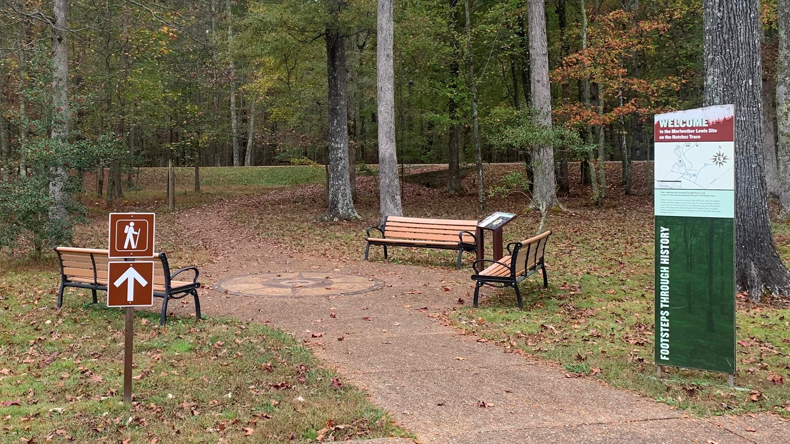 Trailhead with welcome sign on the right. Paved trail leads into the forest away from viewer.