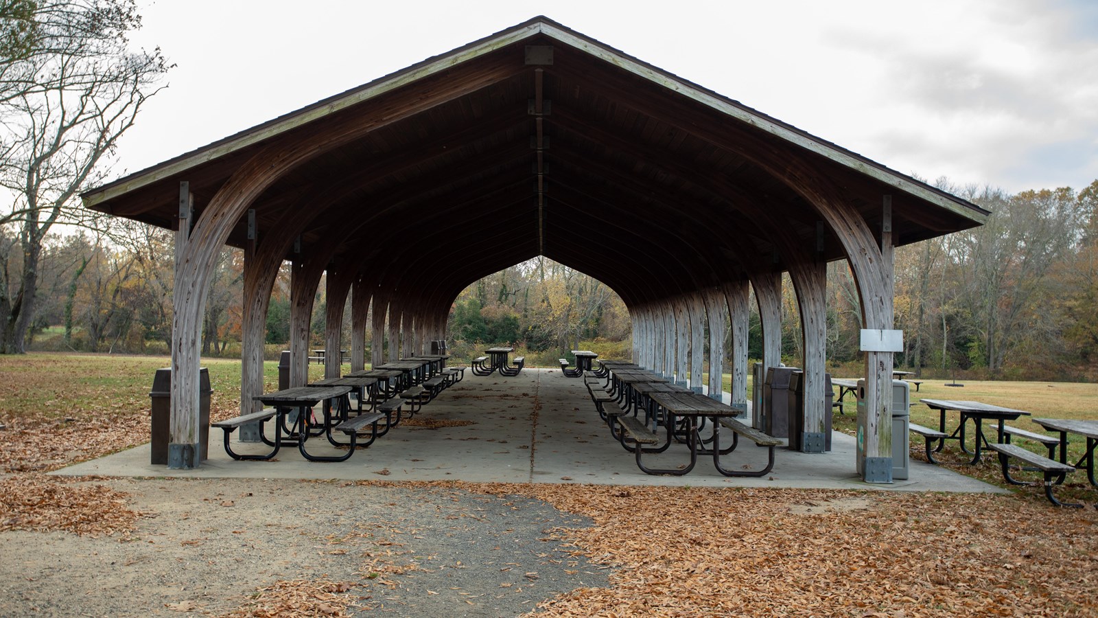 A picnic area with a roof and no walls is in a field. Tables are under and outside the area.