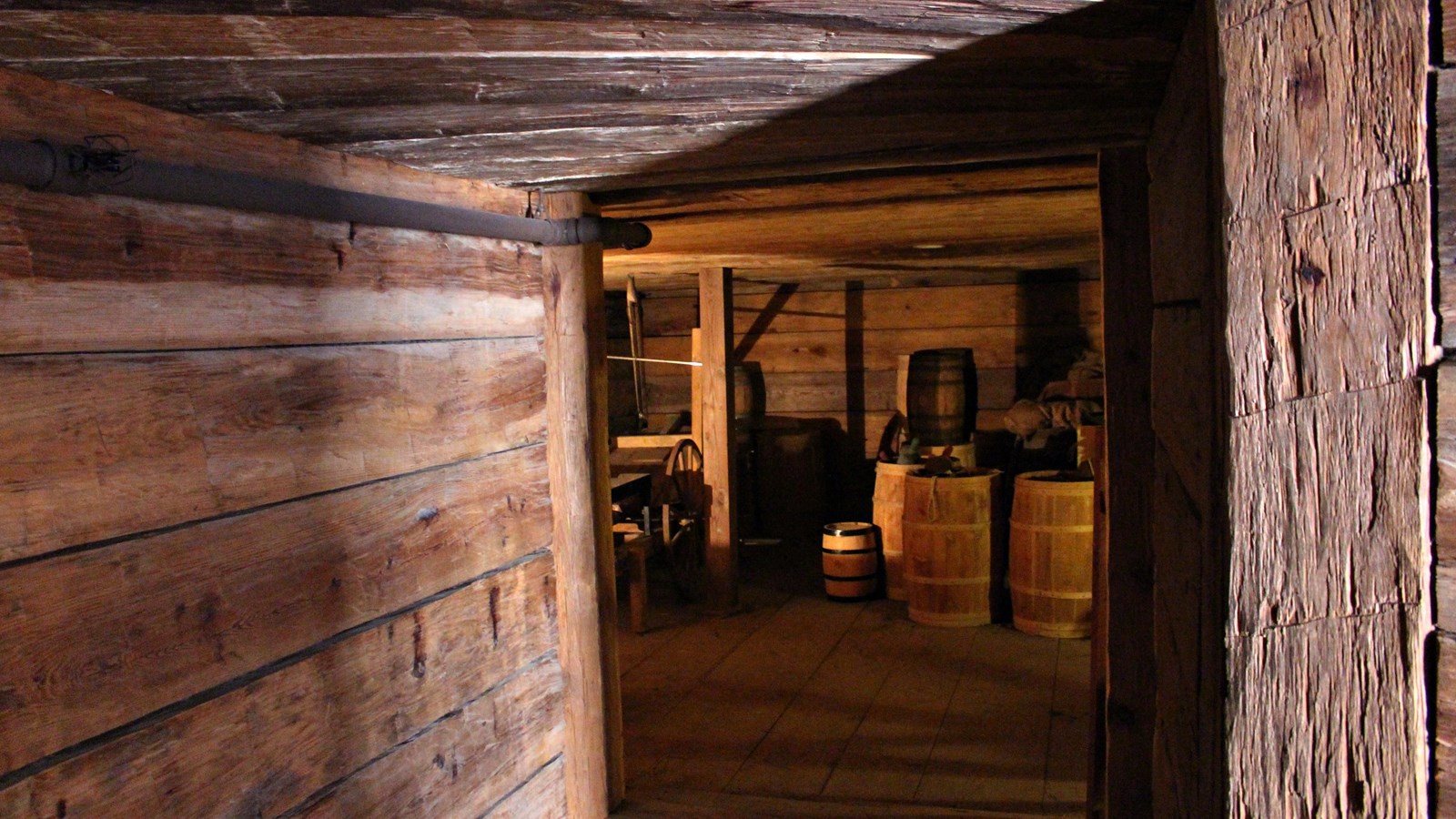Peeking down a tunnel made of angular wood walls, into an larger space with barrels inside. 