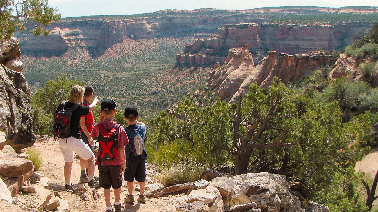 two adults and two children stand on dirt path looking out towards wide open canyons