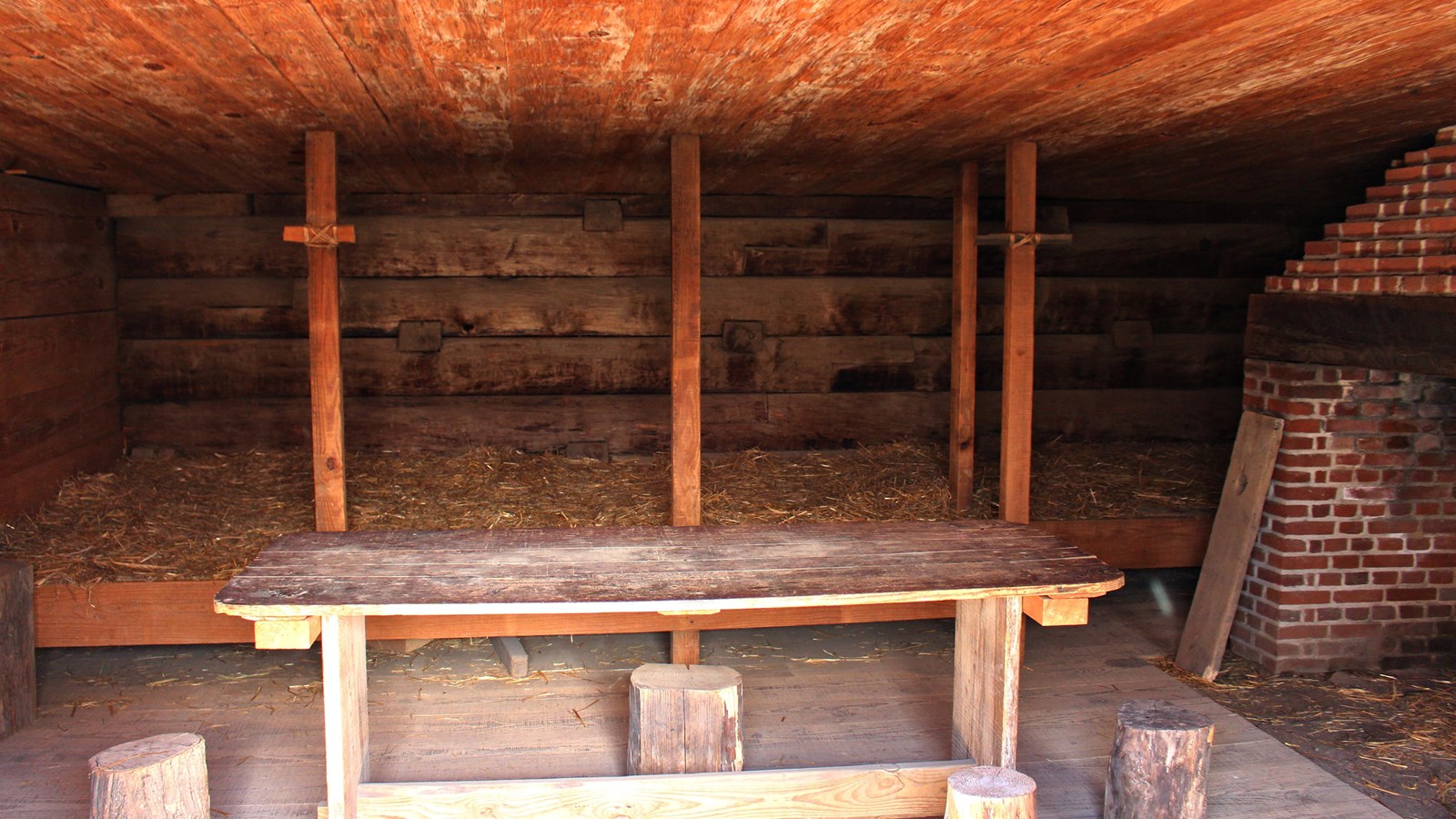 A wooden room with a large fireplace, a big rough wood table, and a bed filled with straw. 
