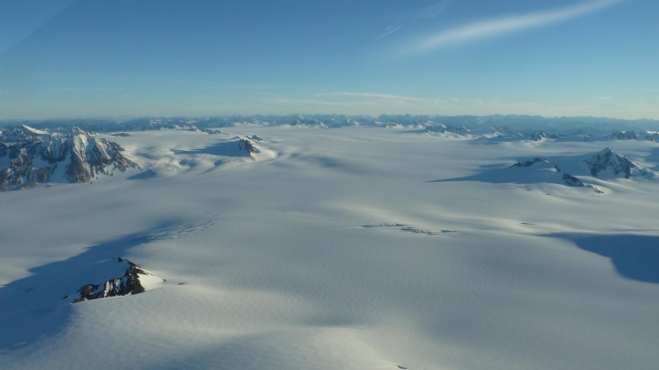 An aerial view of a glacier. Snow covers the land, and mountain tops appear from underneath the ice