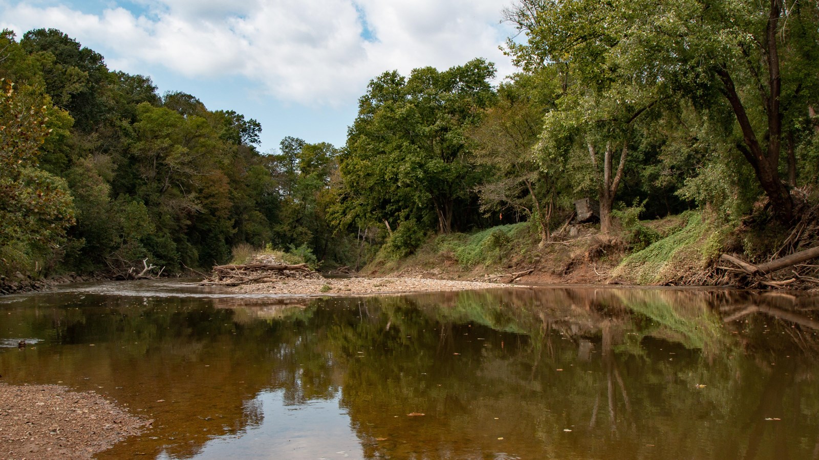 A still section of river reflects the surrounding lush banks under a partially cloudy sky.