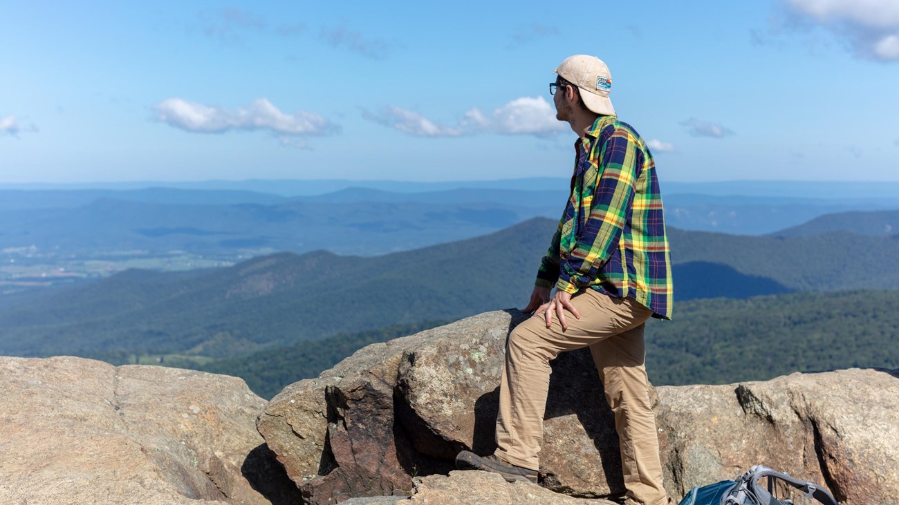 A man in a green shirt and backwards ball cap gazes out over a valley below from a mountain top.