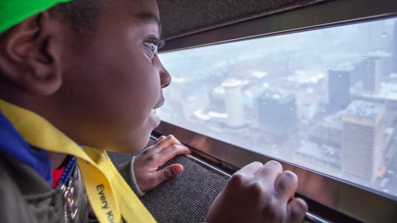 a young African-American girl looks out of the window of the Arch onto the city below