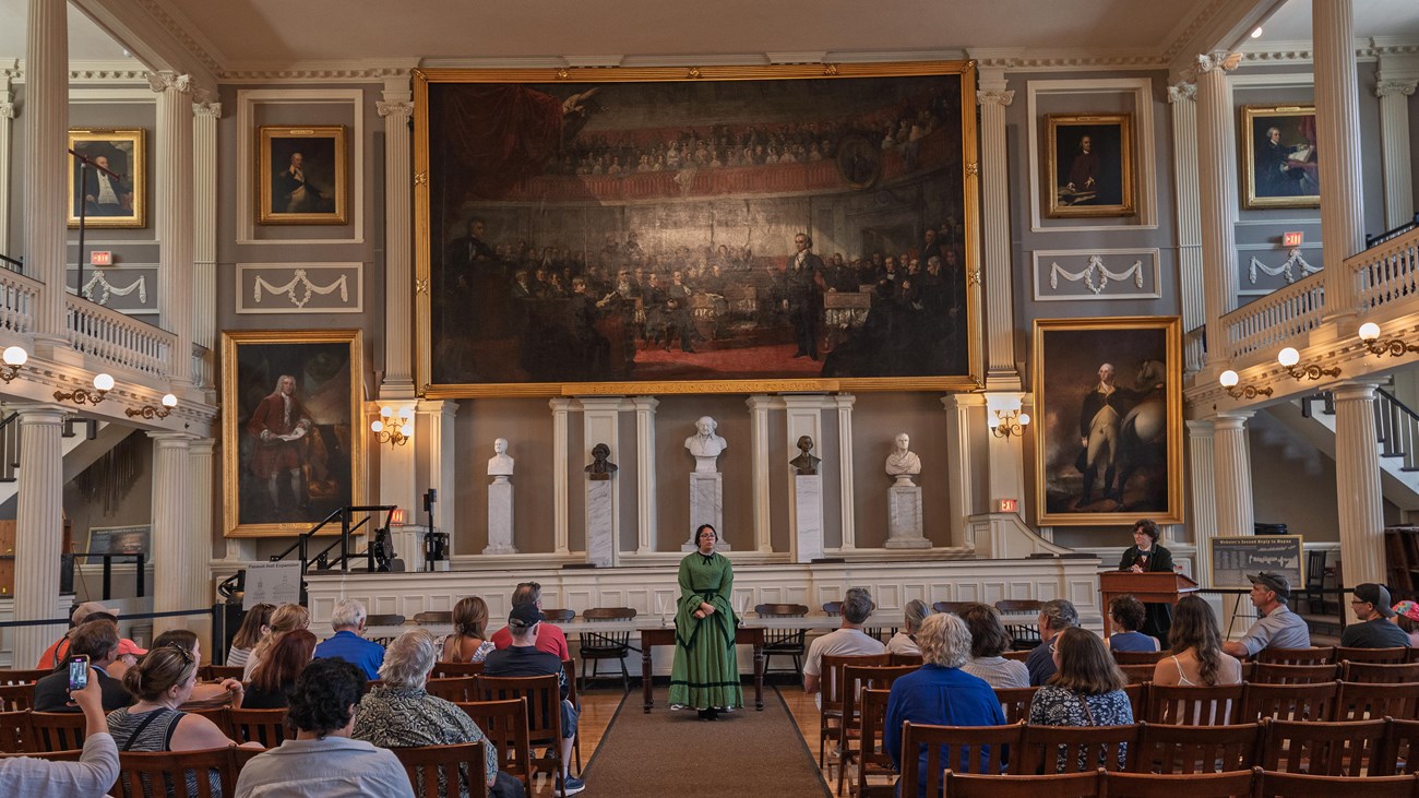 A woman in a green 1800s dress stands in front of the stage and paintings in the Great Hall