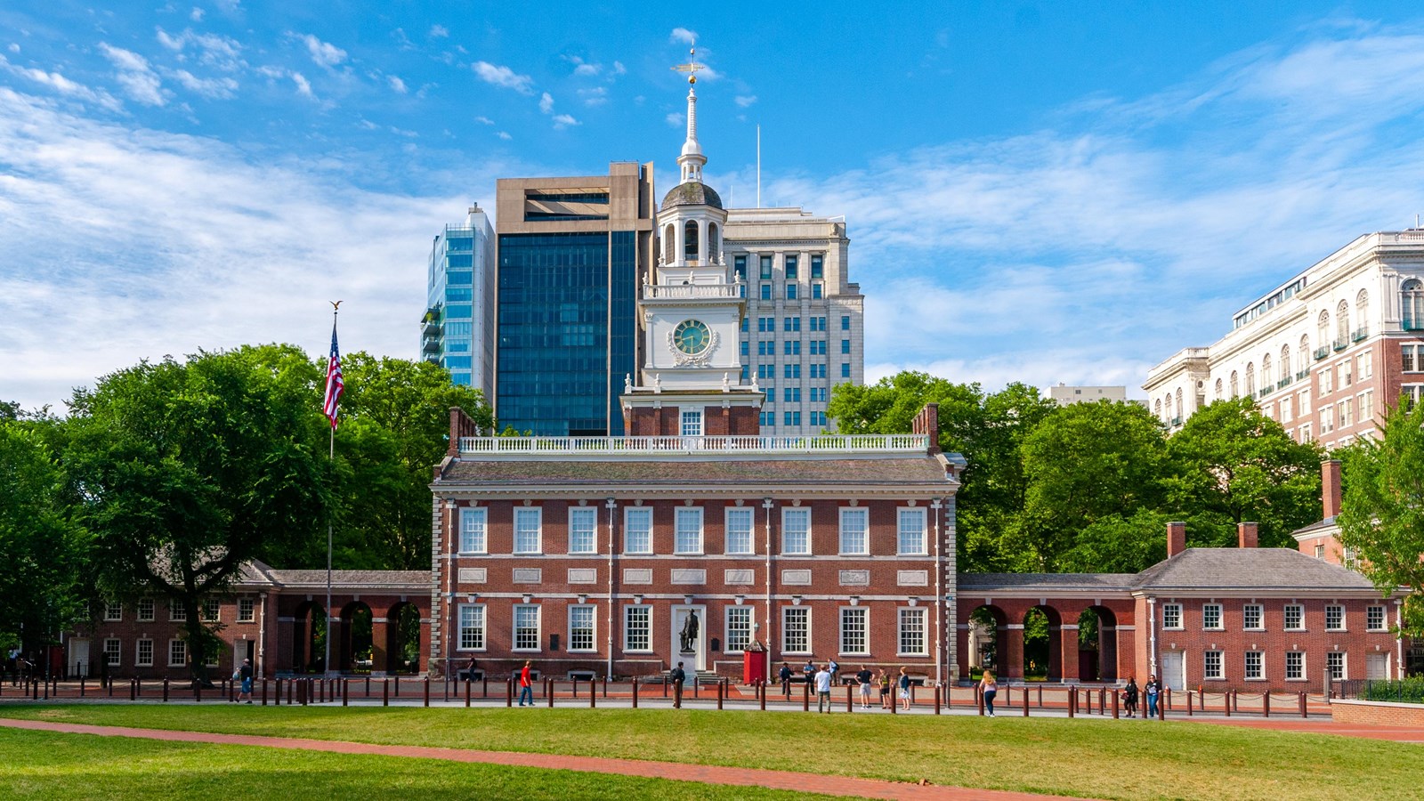 A two story 18th-century brick building with a white steeple surrounded by modern buildings.