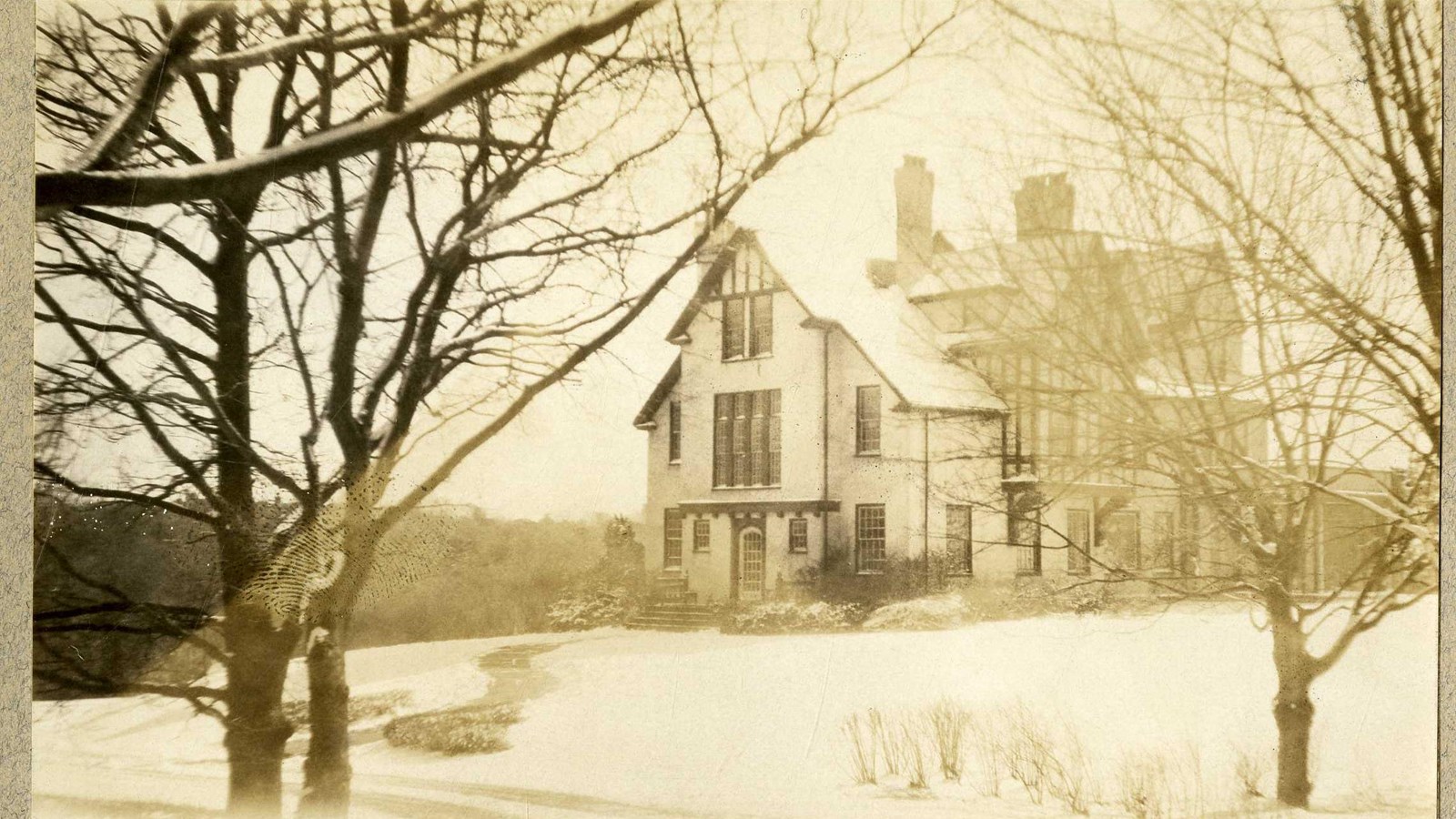 Black and white of large home on flat area with snow and trees all behind, some in front