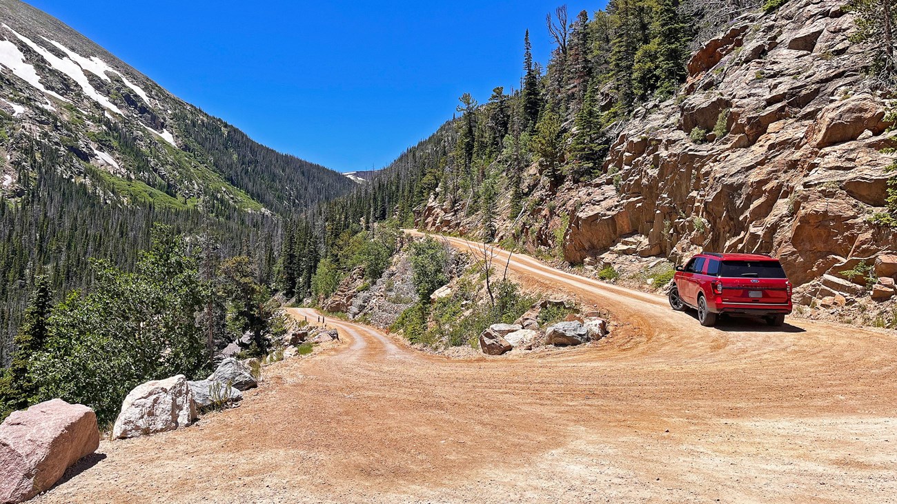 A small SUV is driving on a narrow, dirt road on with switchbacks and view of mountain peaks.