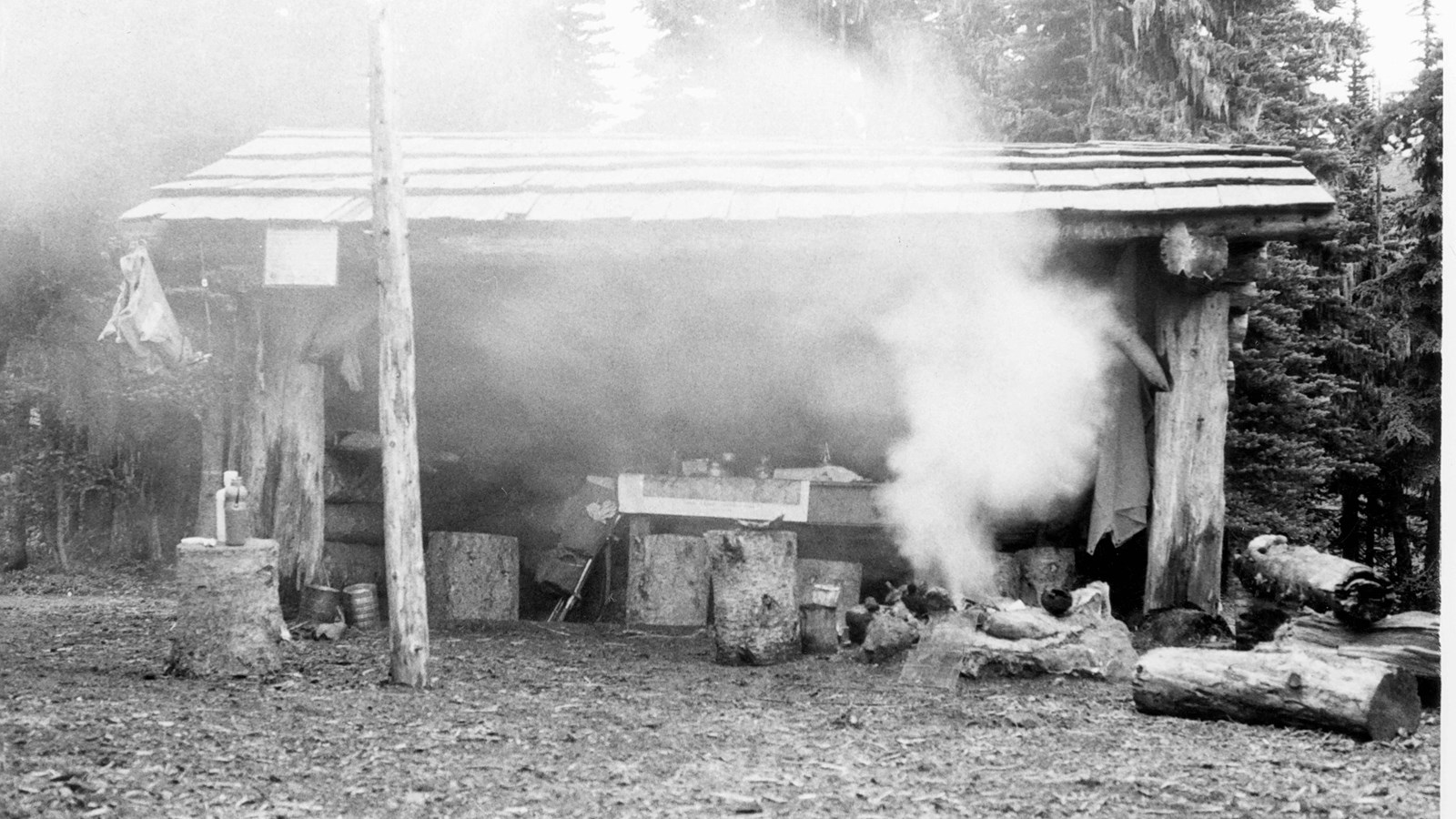 Wooden shelter surrounded by trees with campfire in front of it