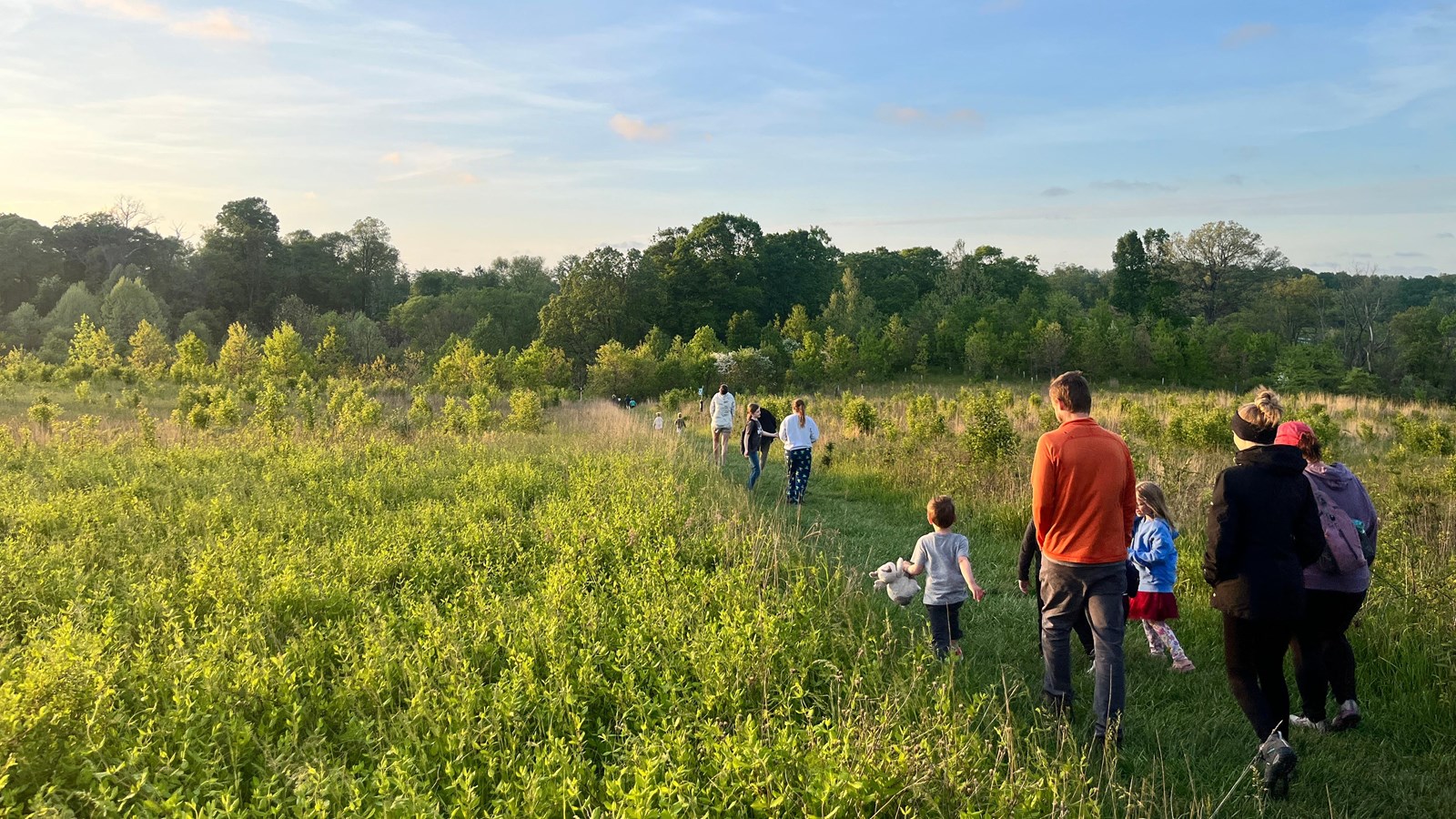 A group of people walking along a grassy path with fields and trees under a blue sky.