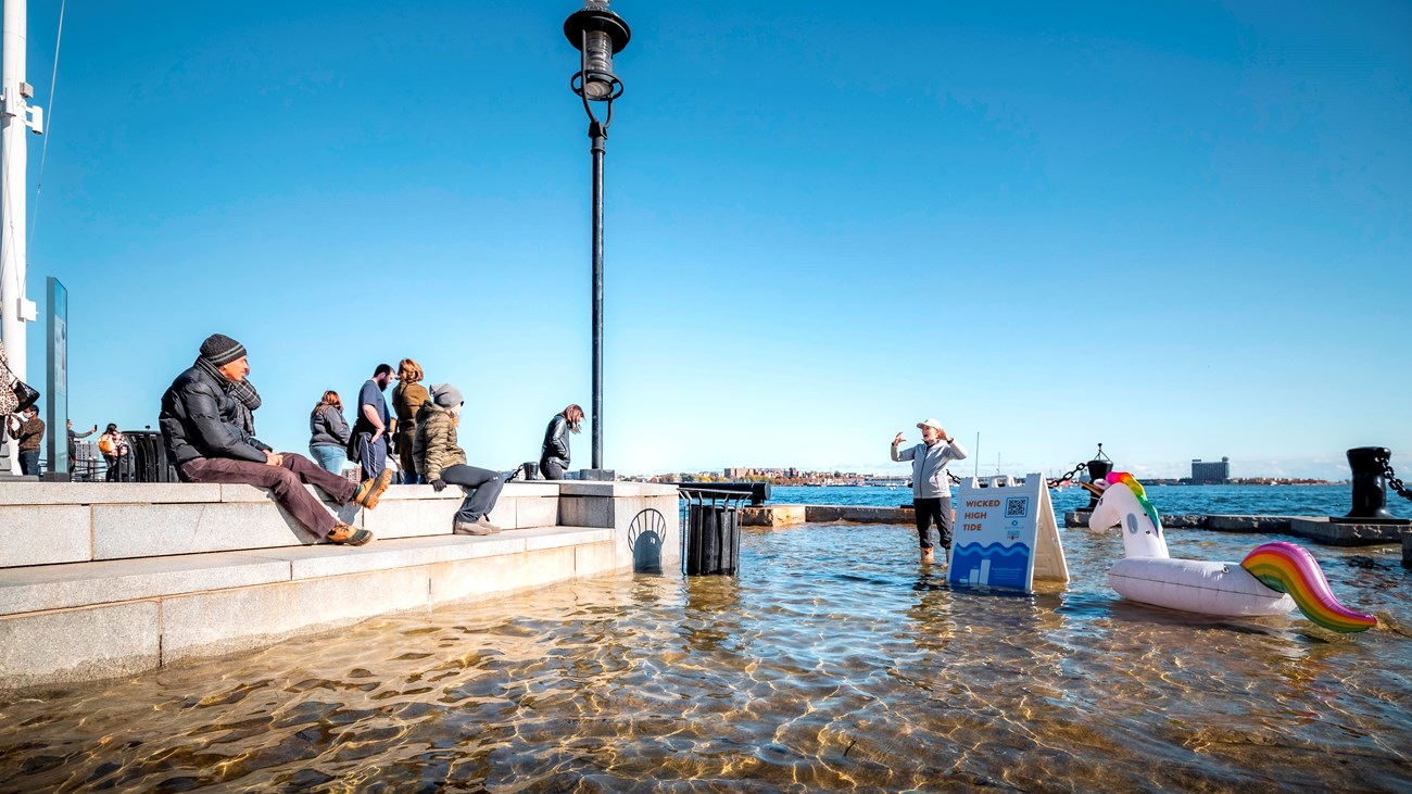 People sitting on Long Wharf as someone in wellies stands in the flooded lower area of the walkway.