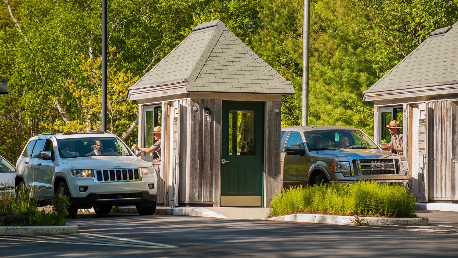 Cars stopped at two small wooden structures in a roadway with uniformed rangers standing at windows.