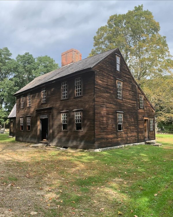 A salt box style colonial home made of wood stands in a grassy field surrounded by trees.