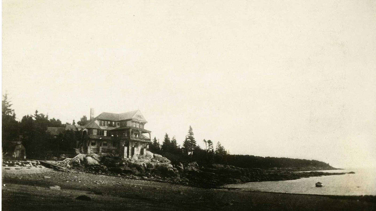 Black and white of large home on beach with trees around it and rocks below it by the water