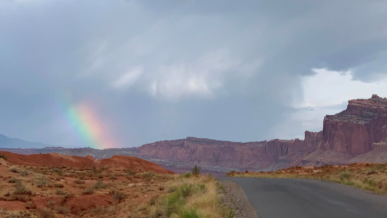 Stormy skies with a small spot of a rainbow, red cliffs, and a blacktop road.