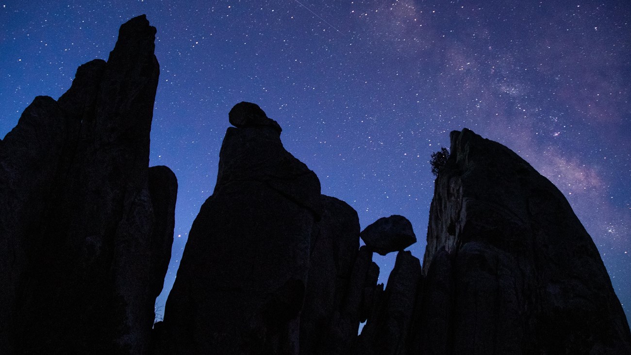 Silhouettes of 3 large pinnacles and a smaller balanced boulder under a purple and blue starry sky.