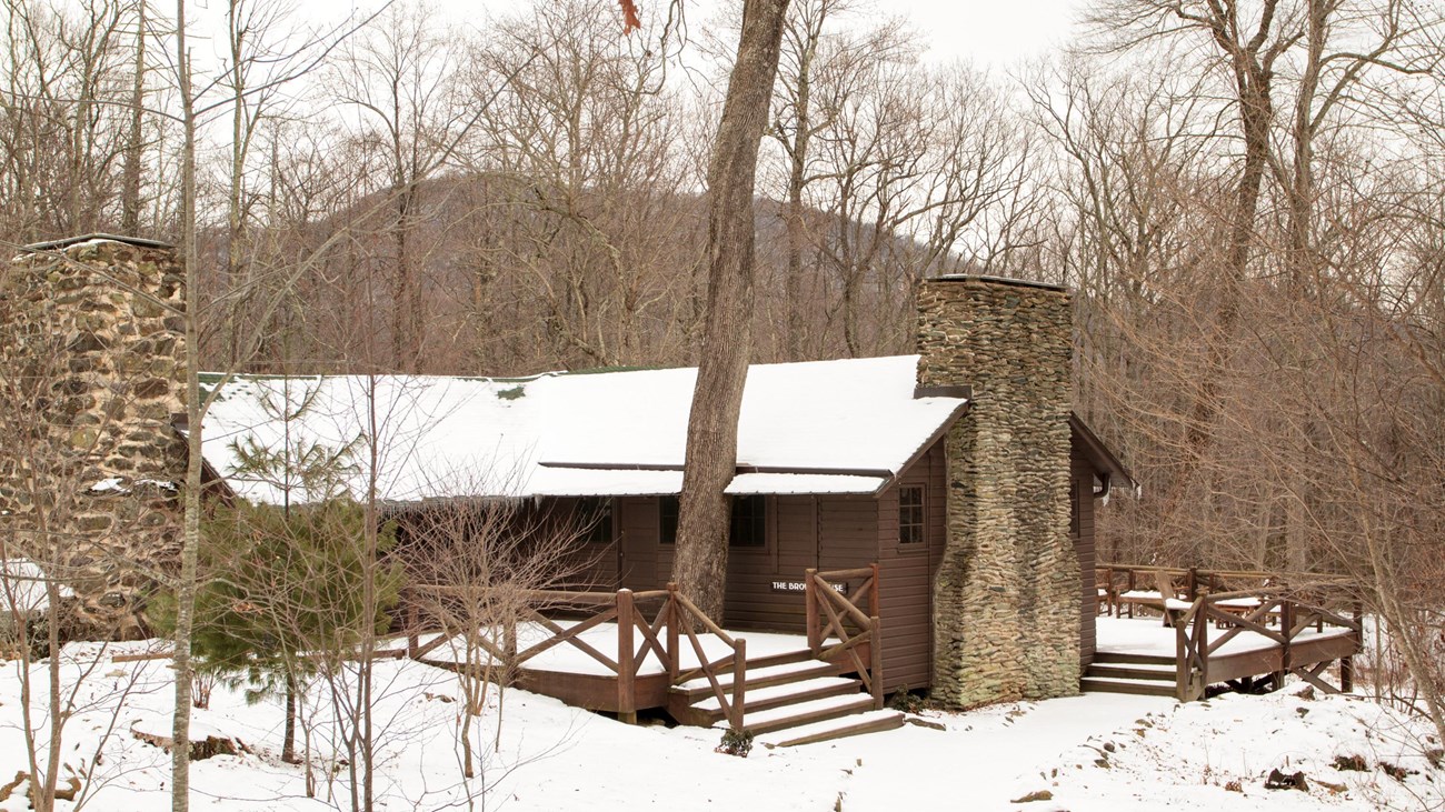 An historic cabin with a large porch sits in the middle of a snow-covered forest.
