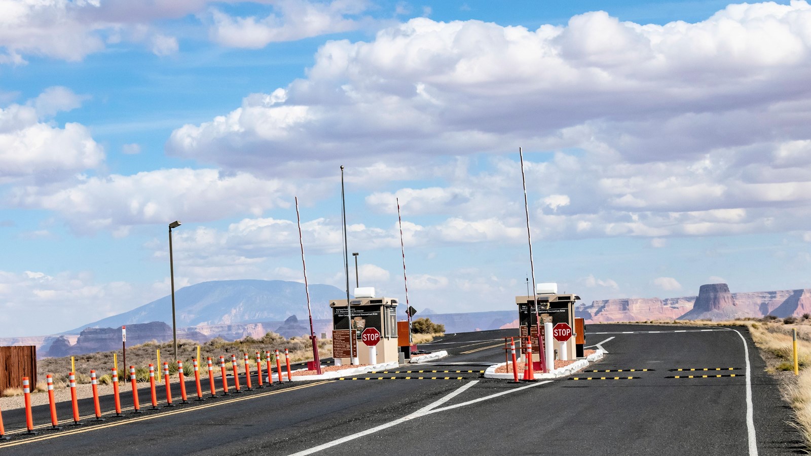 Asphalt road with two entrance lanes that lead to two beige booths, orange cones, barricades, and st