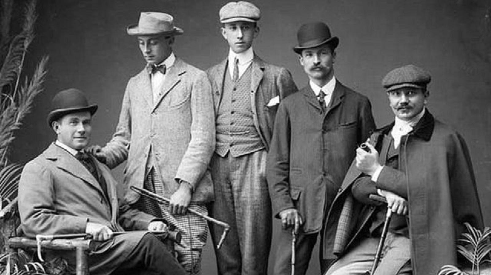 Black and white photo of five men in 1900. The two on each end are seated, all don hat and cane.all 