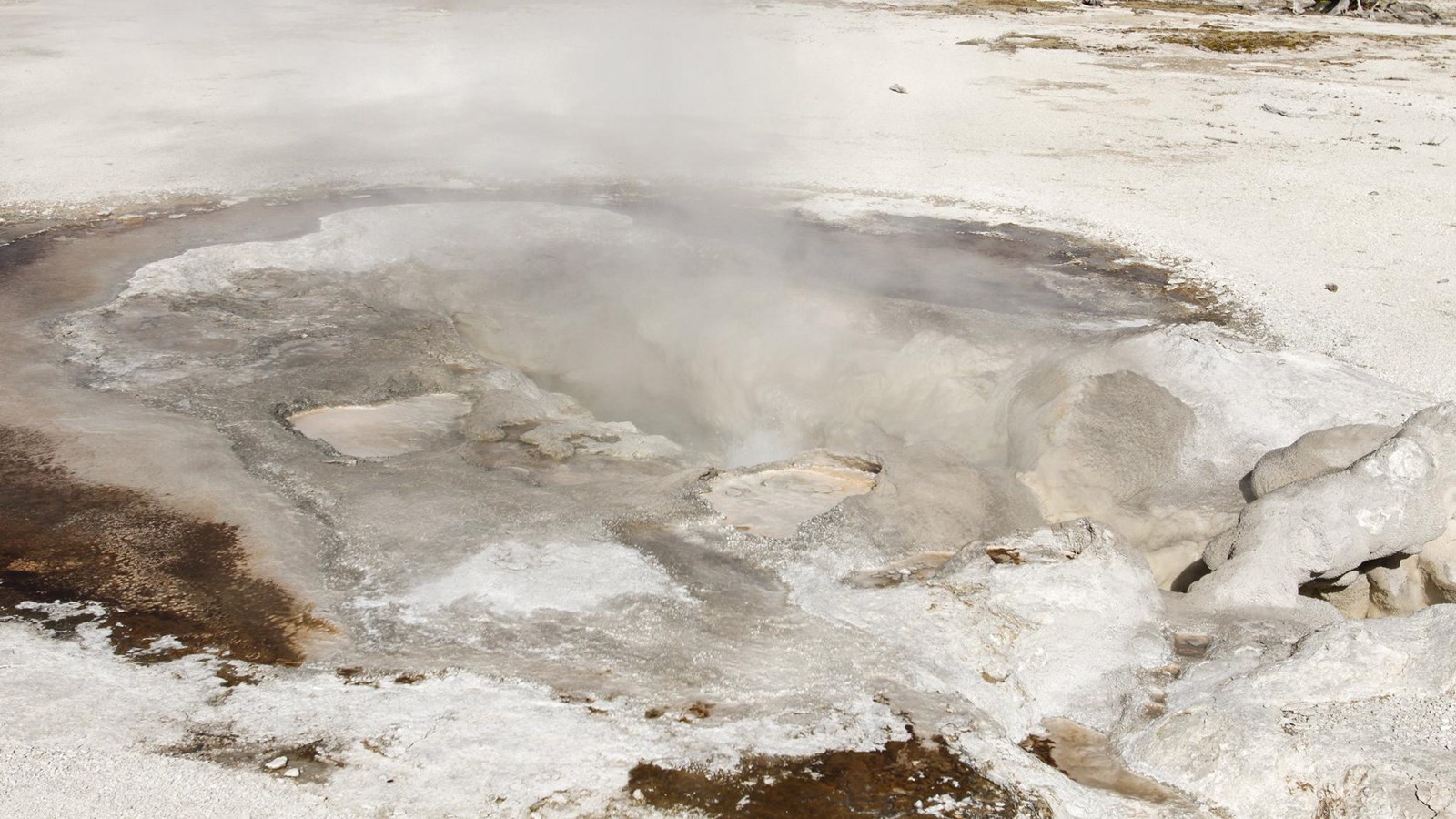 Steam and water escape from a thermal vent located on barren ground in a thermal basin.