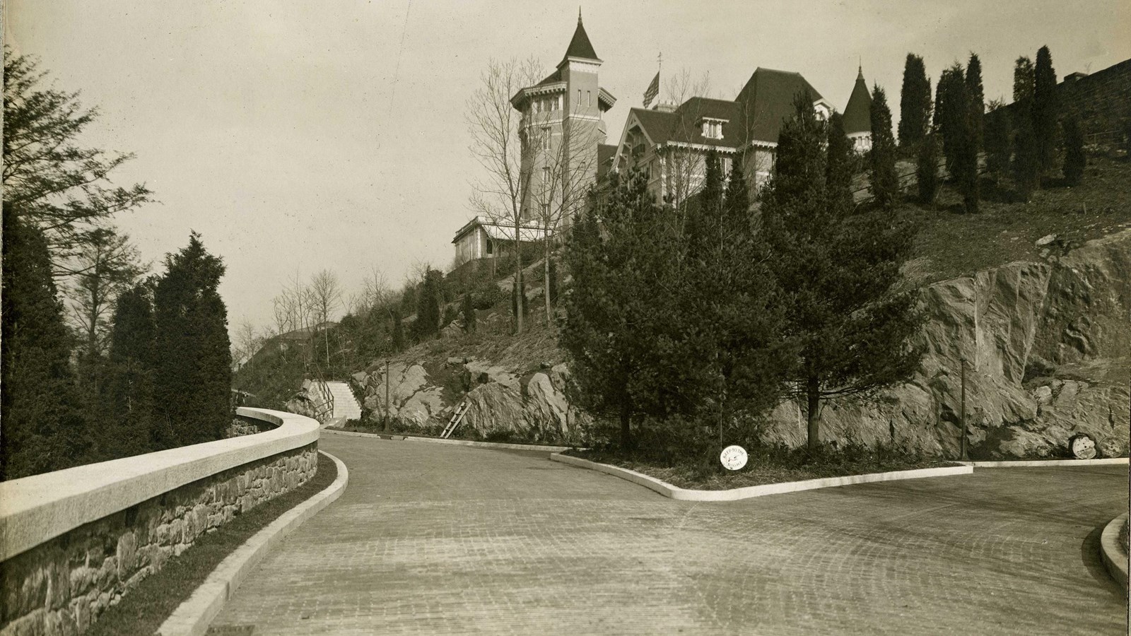 Black and white of road lined with stone wall and rocky cliff with building on top lined with trees