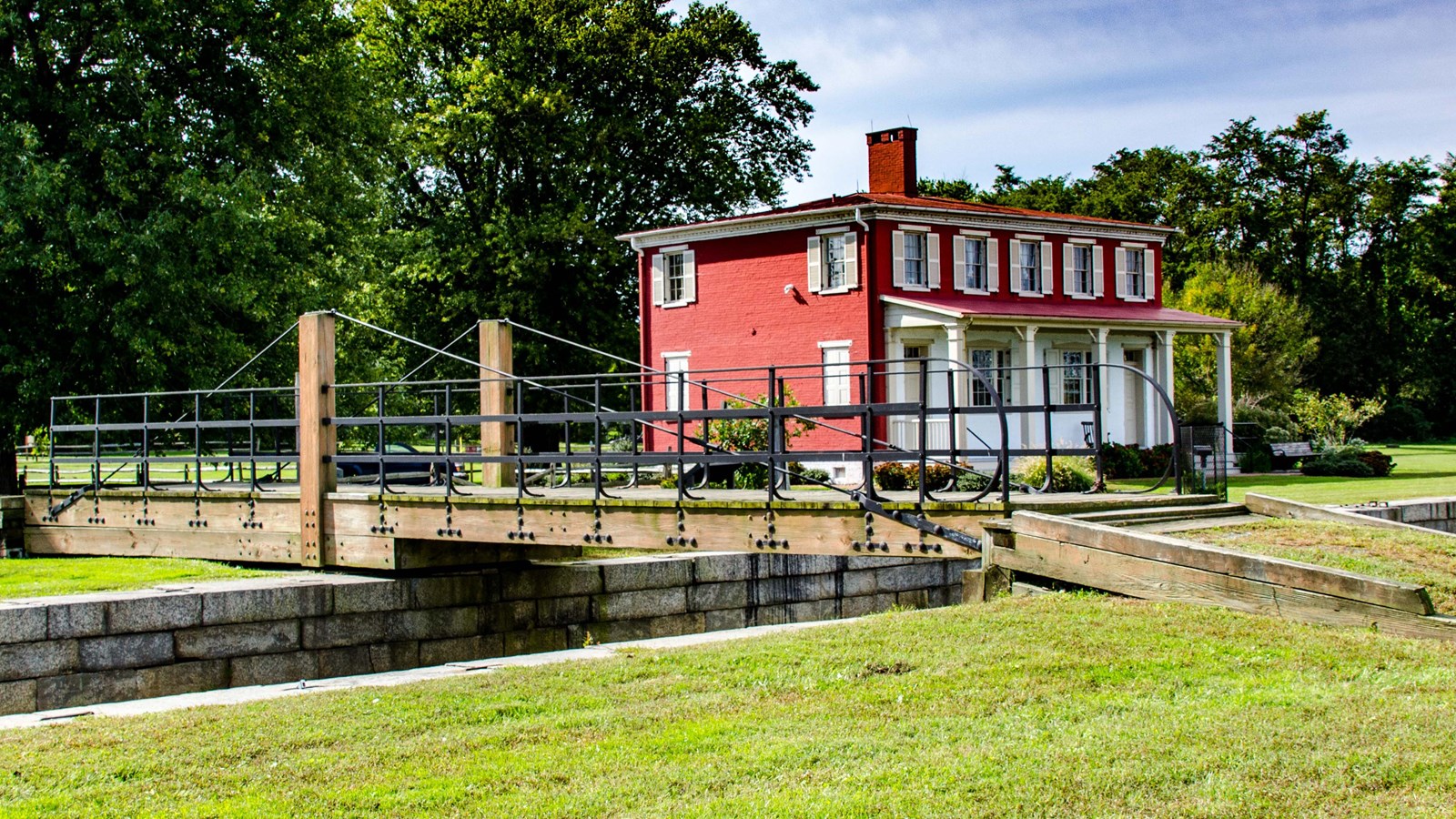 A red brick two-story building with a small wooden bridge over a stone canal, surrounded by greenery