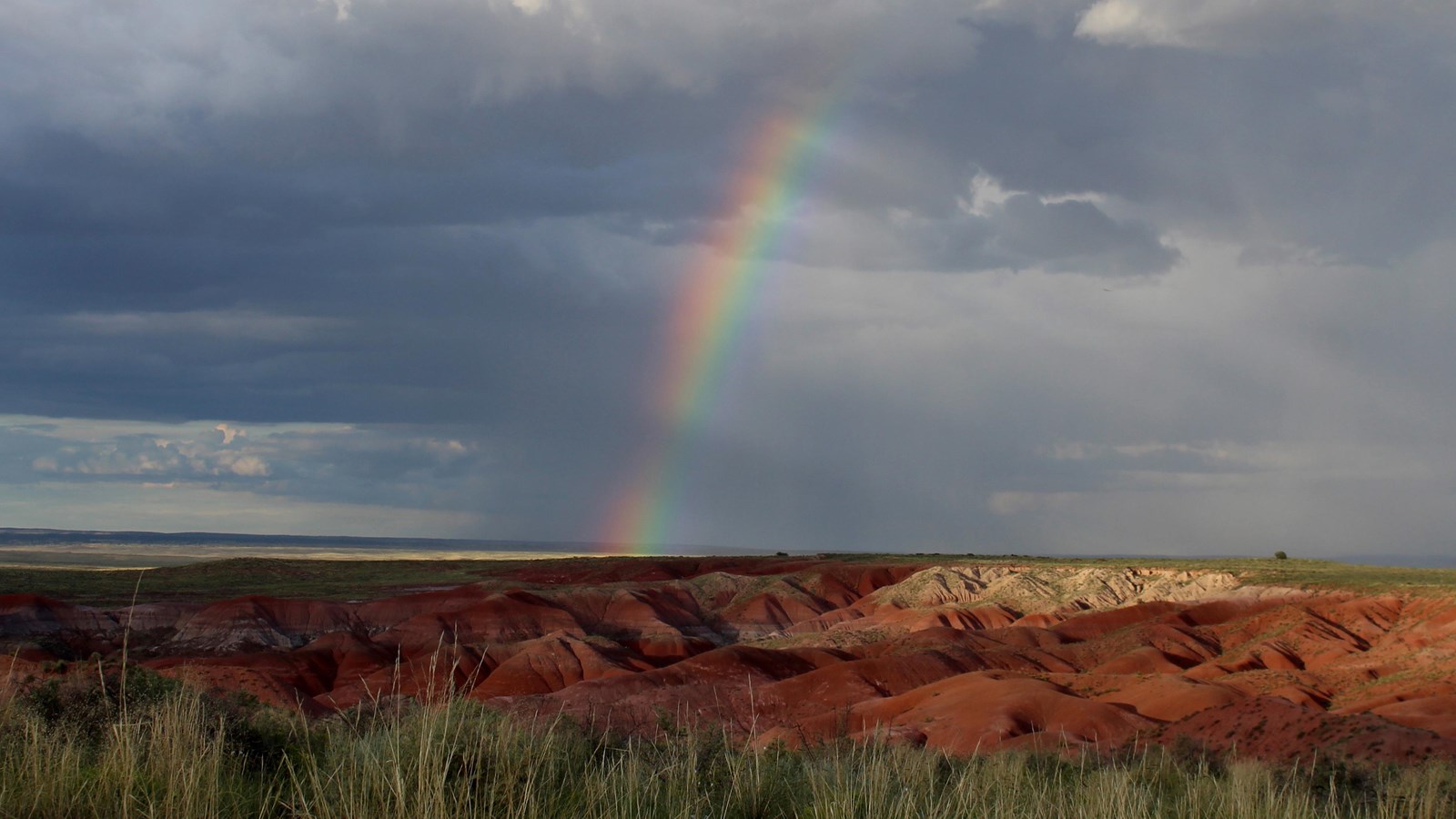 Rainbow against a cloudy sky above red badlands and grassland.