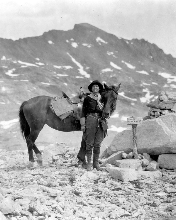 A woman stands next to a horse surrounded by rocks and mountains. A sign nearby says "Muir Pass"