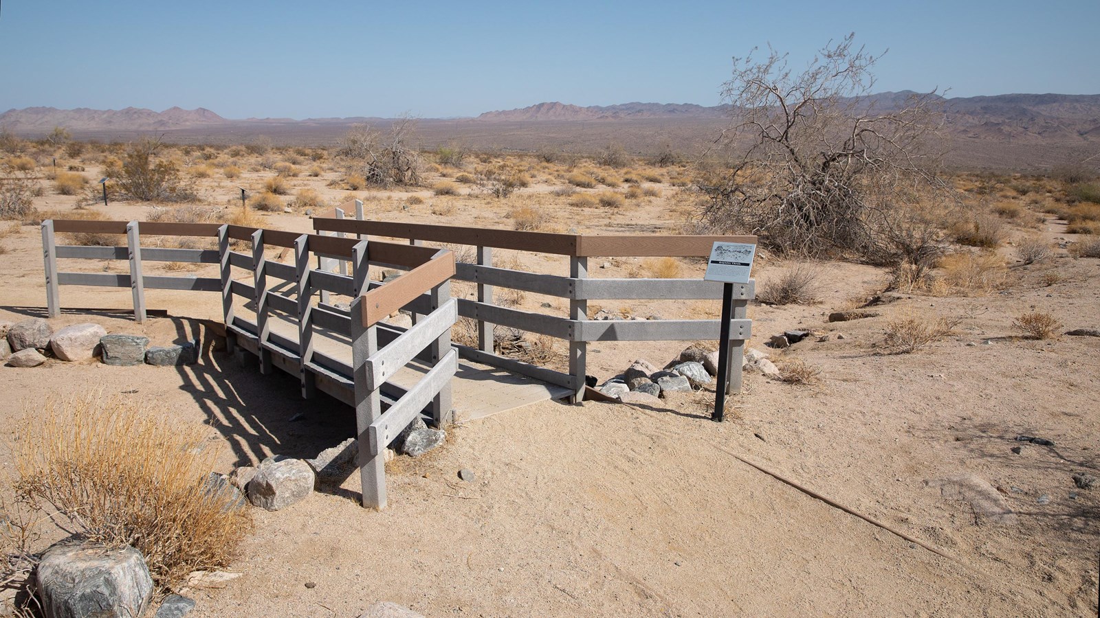 A dirt path and wooden footbridge with scattered shrubs in the area and mountains in the distance.