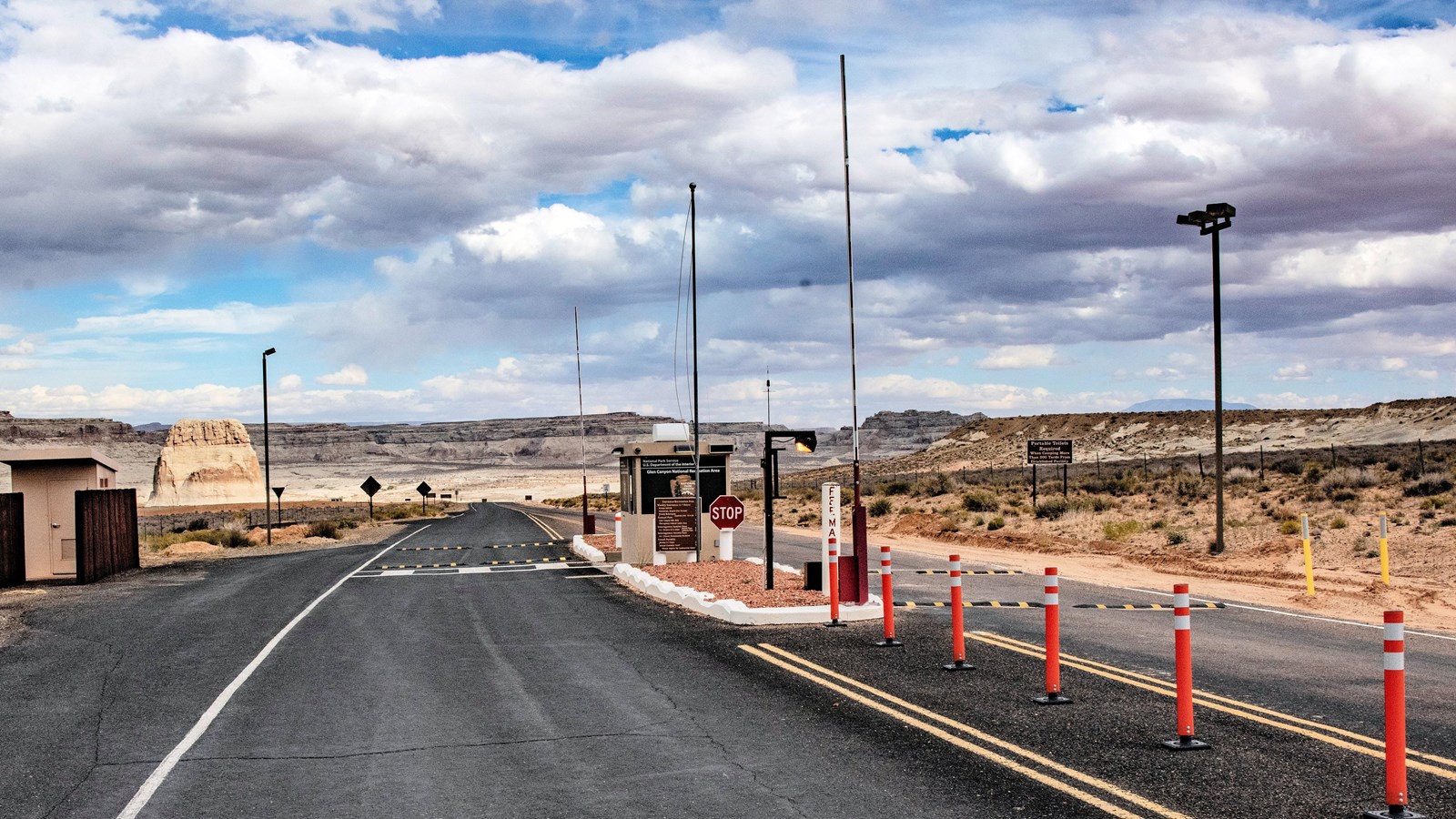 Two lans asphalt road that leads to and beyond a beige metal booth. Lane barricades, orange cones, s