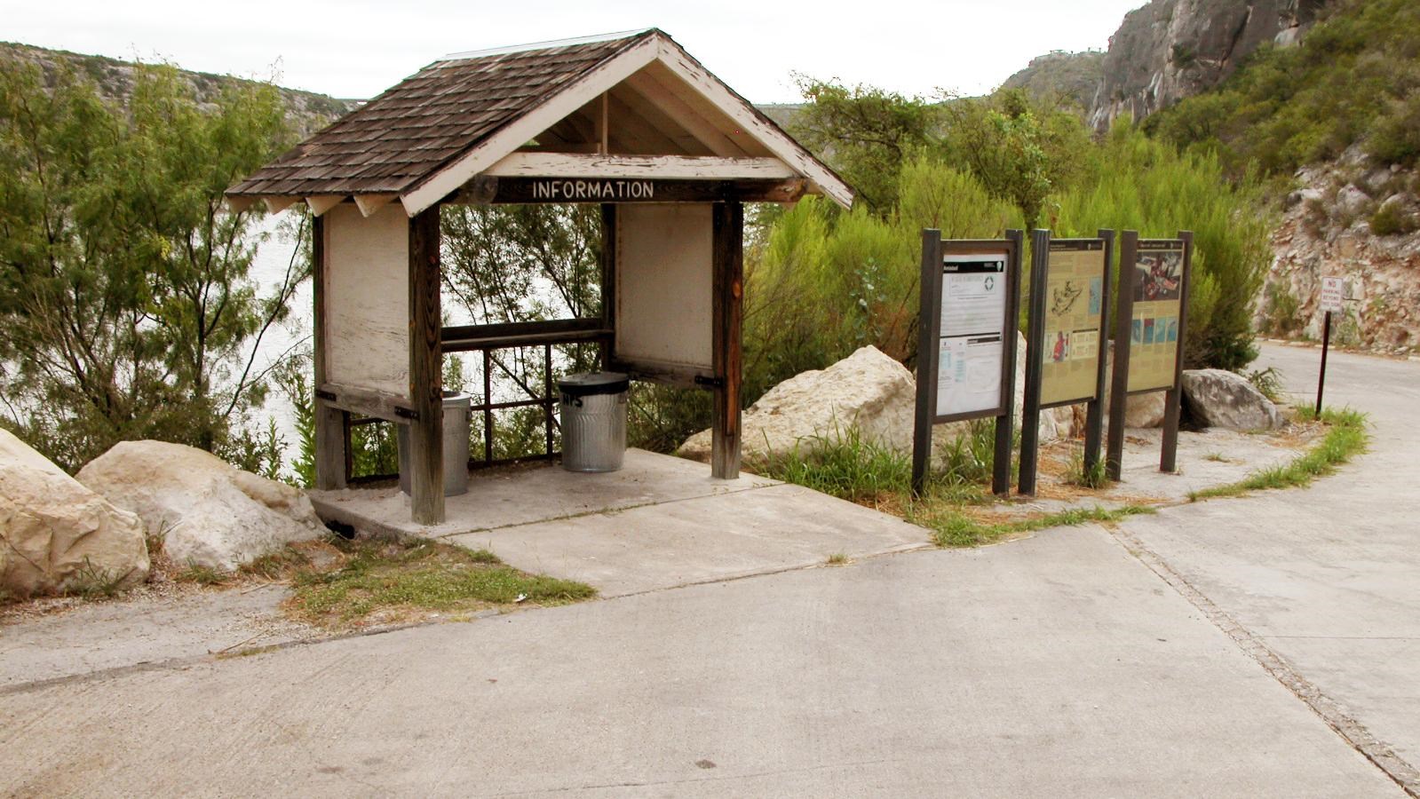 Wooden shade shelter with trash cans inside and 3 information panels next to it.