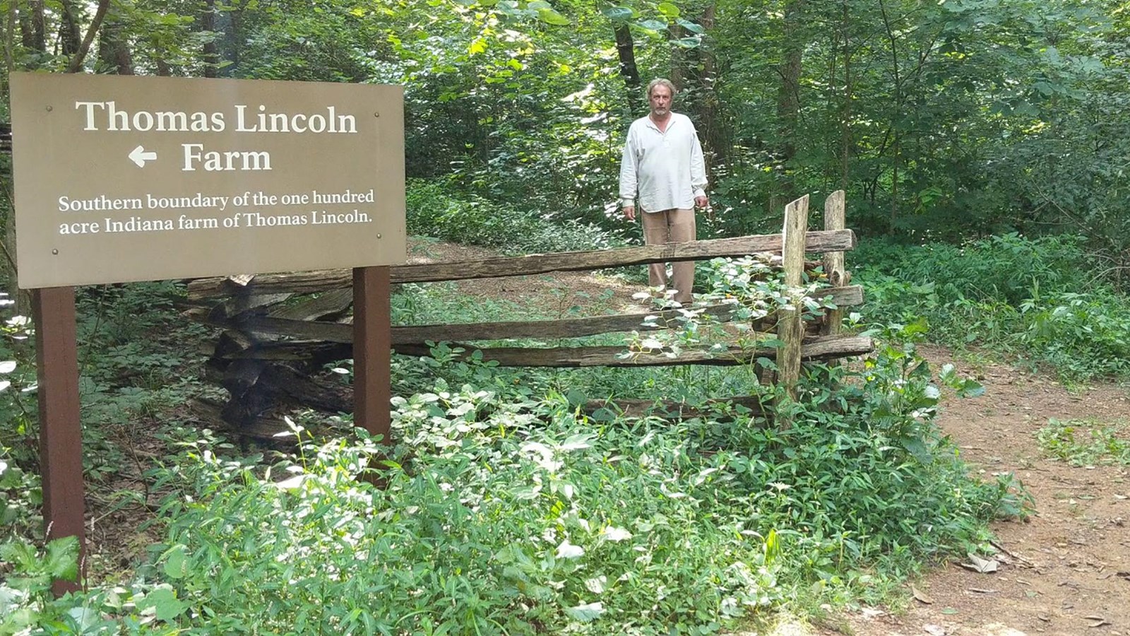 A man stands on a trail next to a split-rail fence. A sign is at the junction of two trails. 