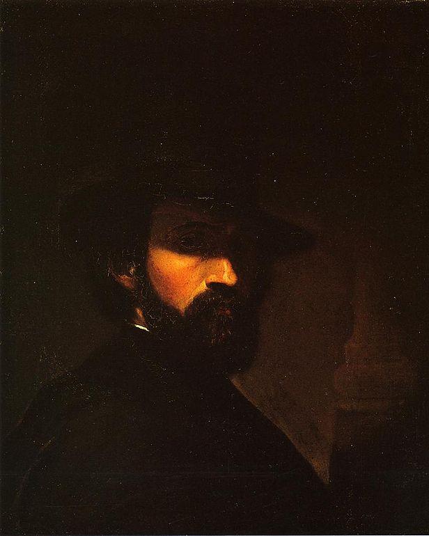 A man with a dark mustache and beard wears a dark hat and suit jacket.