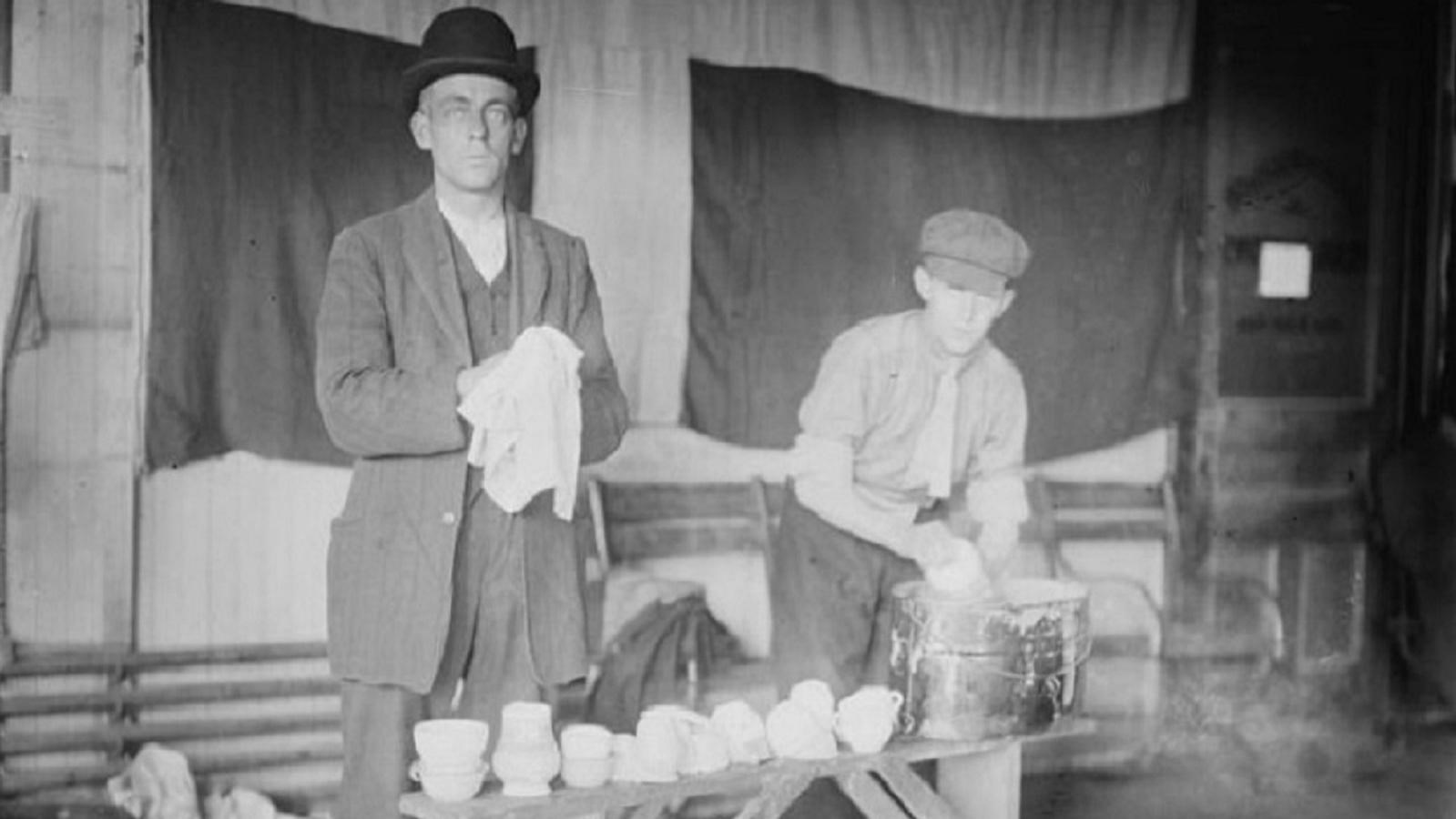 Black and white photo of two men washing dishes in a bucket on a crude wooden table behind a kitchen
