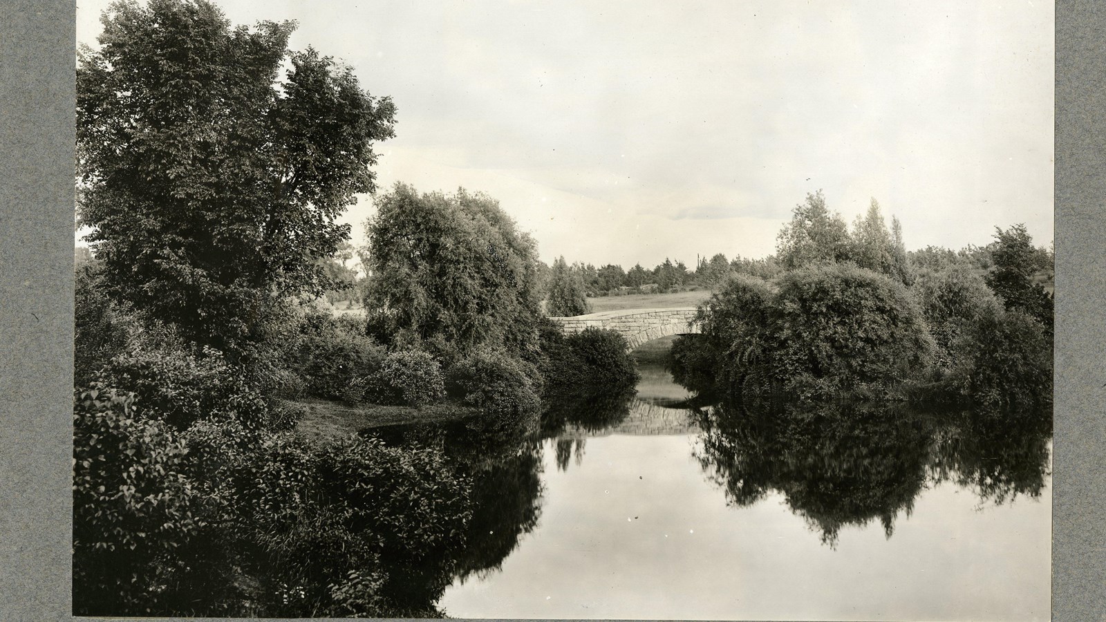 Black and white of stone bridge going over water with shrubs on edge of water