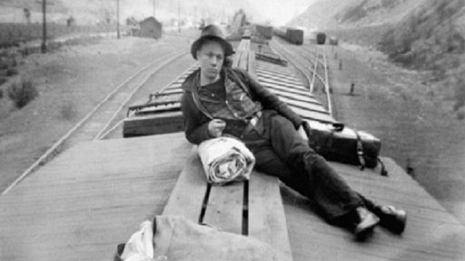 Black and white photo of man riding on top of a train with a bedroll. 